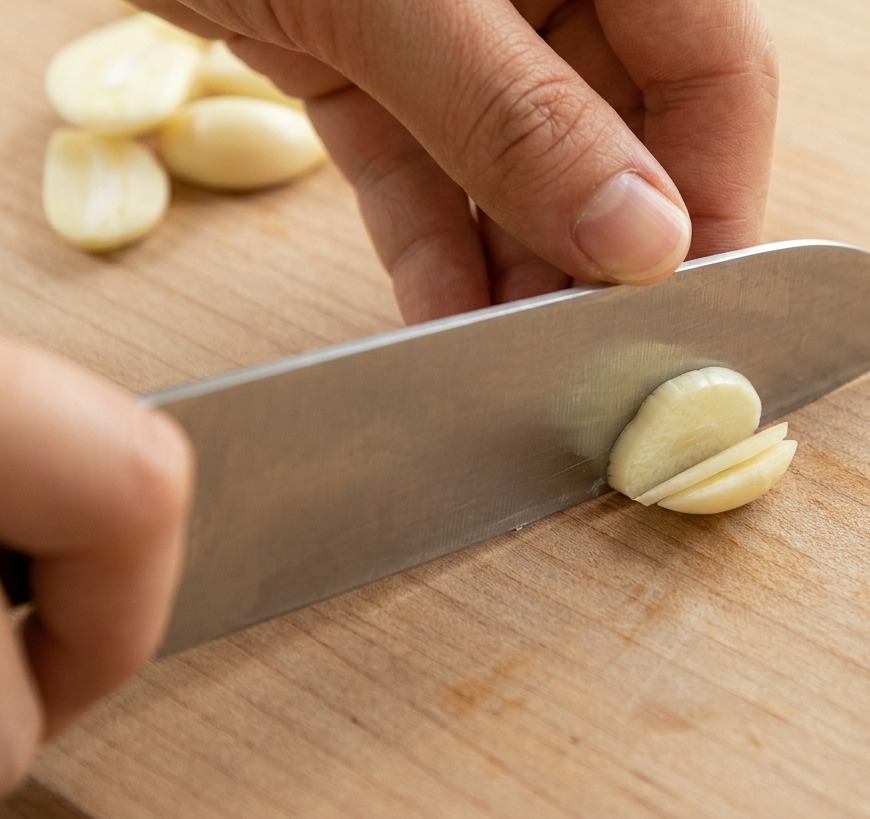 A close-up shot of hands using a sharp knife to slice whole garlic cloves into thin rounds on a wooden cutting board.