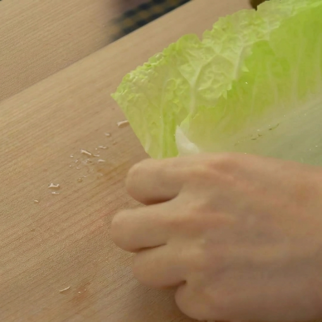 Hands slicing through a stack of pale green napa cabbage leaves to create wide strips on a wooden cutting board.