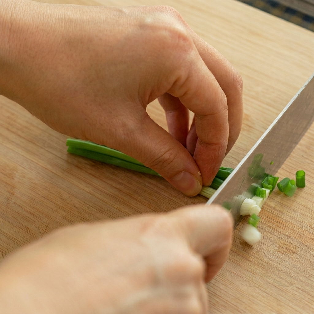 A close-up of a chefs knife finely dicing a bunch of green onion stalks into small green and white rings.