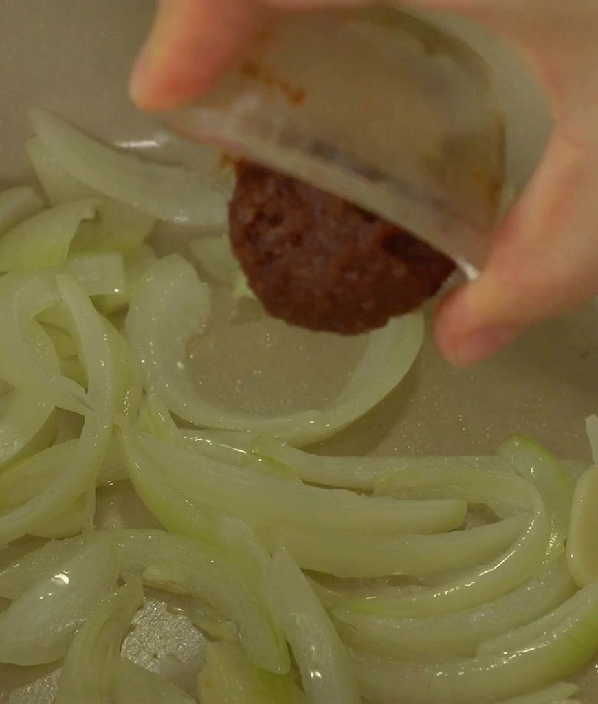 A hand pouring a dollop of dark brown fermented soybean paste from a small bowl into a pot containing translucent sautéed onions.