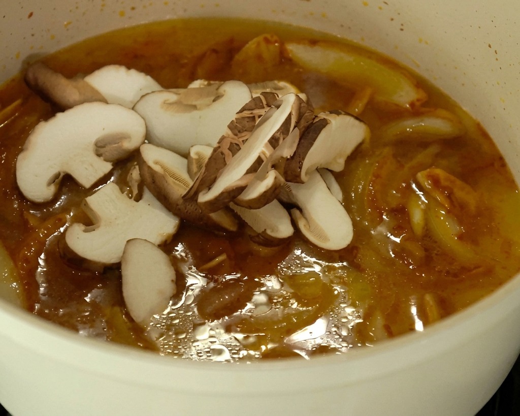 Freshly sliced mushrooms and pieces of cabbage resting on the surface of a rich, orange-colored simmering ramen broth in a white pot.