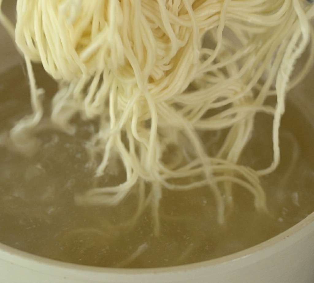 A bundle of thin, pale ramen noodles being submerged into a pot of rapidly boiling water.