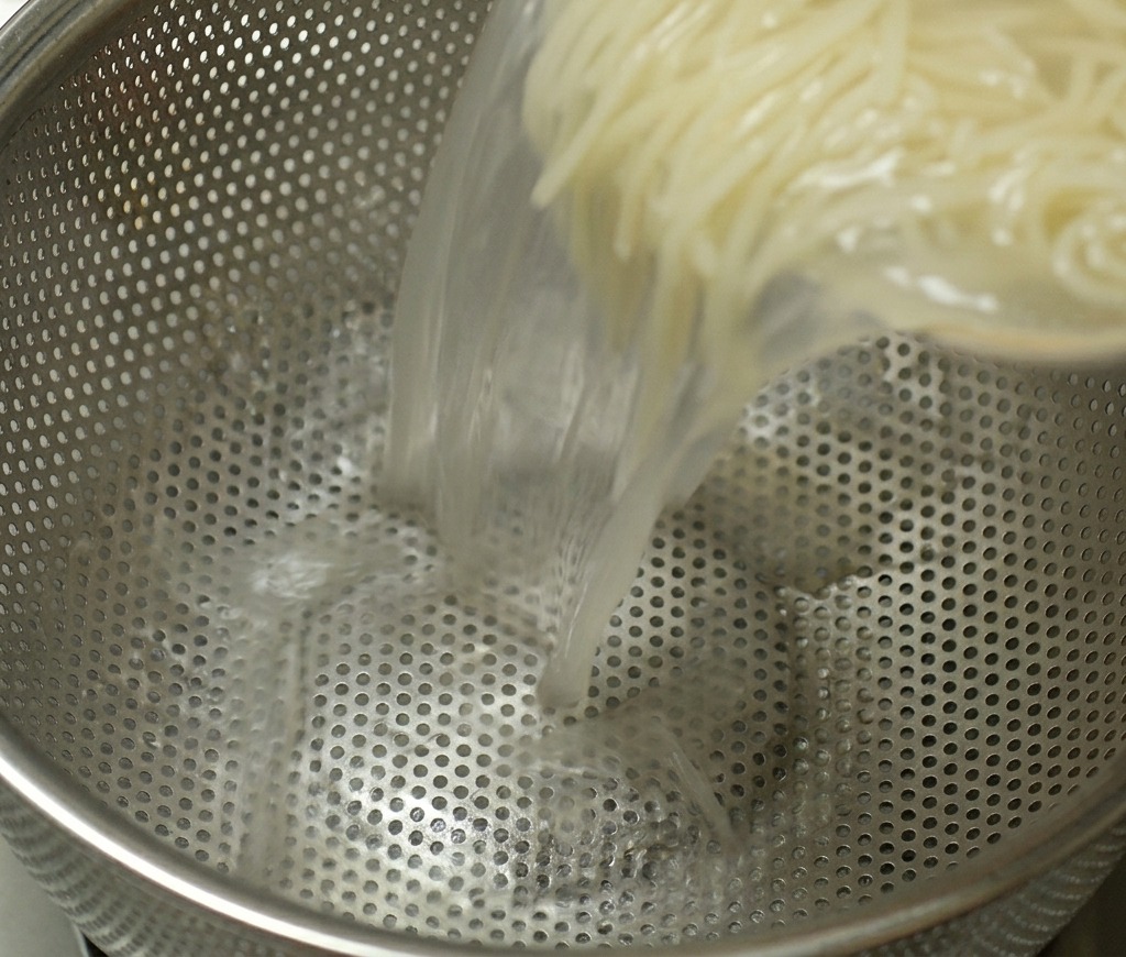 A pot of cooked ramen noodles being poured into a stainless steel colander to drain the hot cooking liquid.