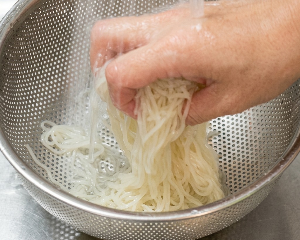A hand tossing white ramen noodles in a stainless steel colander under a stream of cold water in a sink.