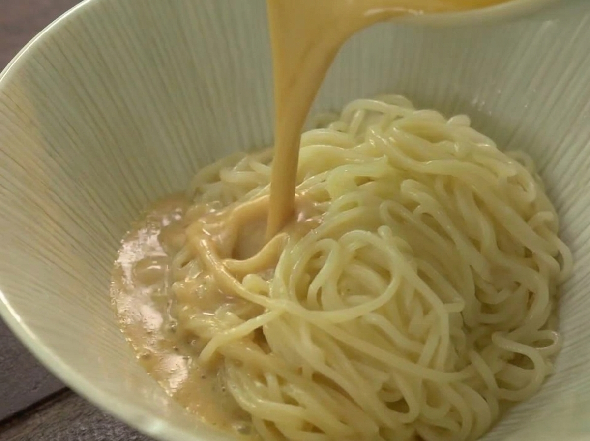 A stream of hot, creamy beige ramen broth being poured over a mound of noodles in a light-colored ceramic bowl.