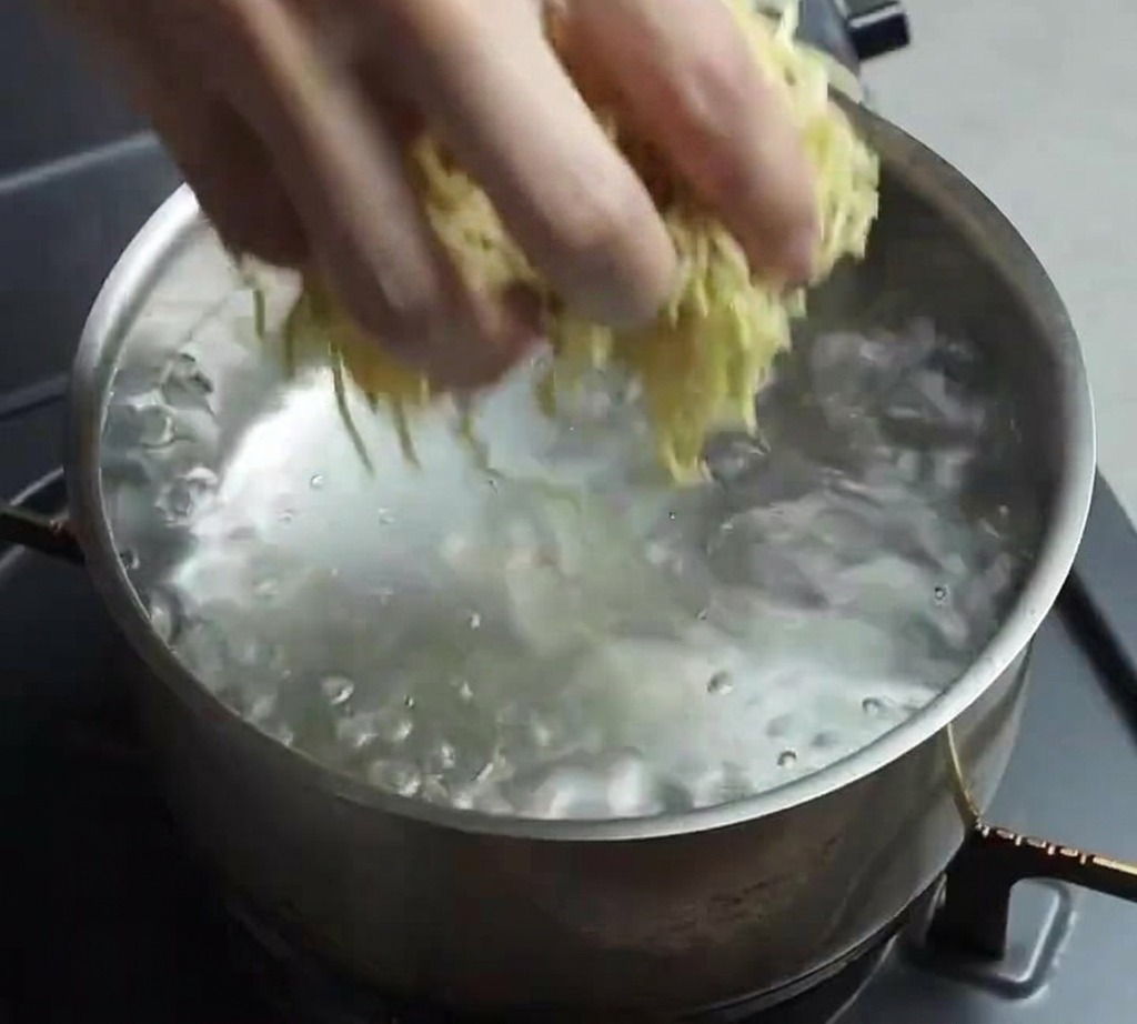 A hand dropping a bundle of fresh yellow ramen noodles into a stainless steel pot of bubbling boiling water.