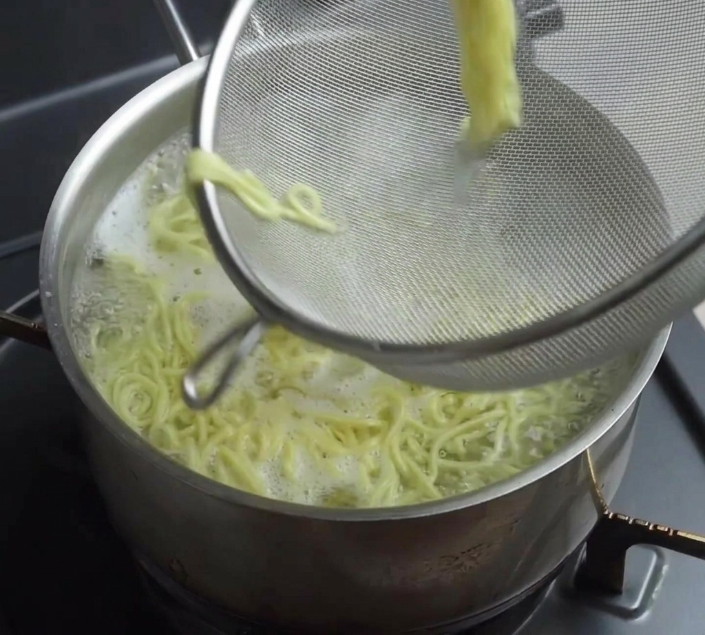 A wire mesh strainer lifting a portion of cooked yellow ramen noodles out of a steaming pot.