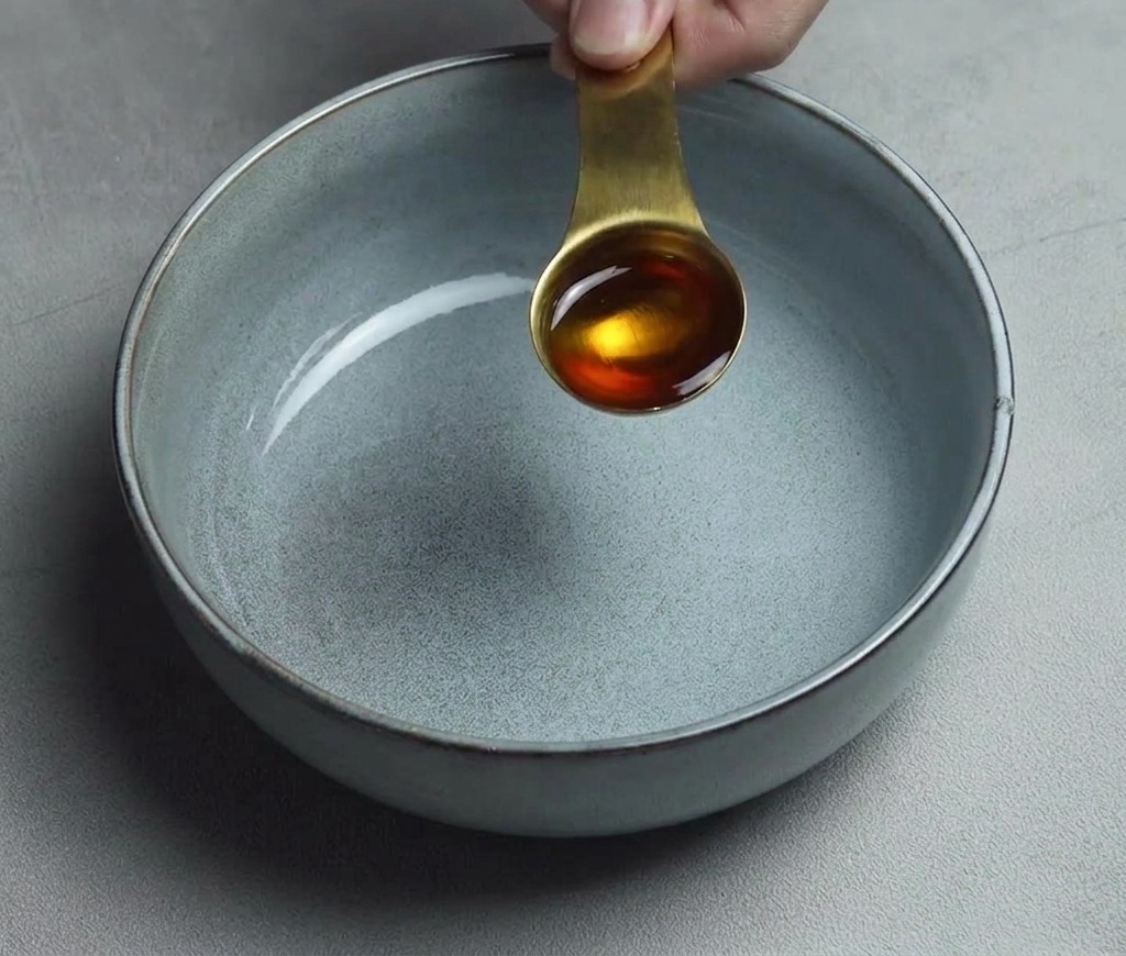 A golden tablespoon of sesame oil being poured into a clean, empty gray ceramic bowl.