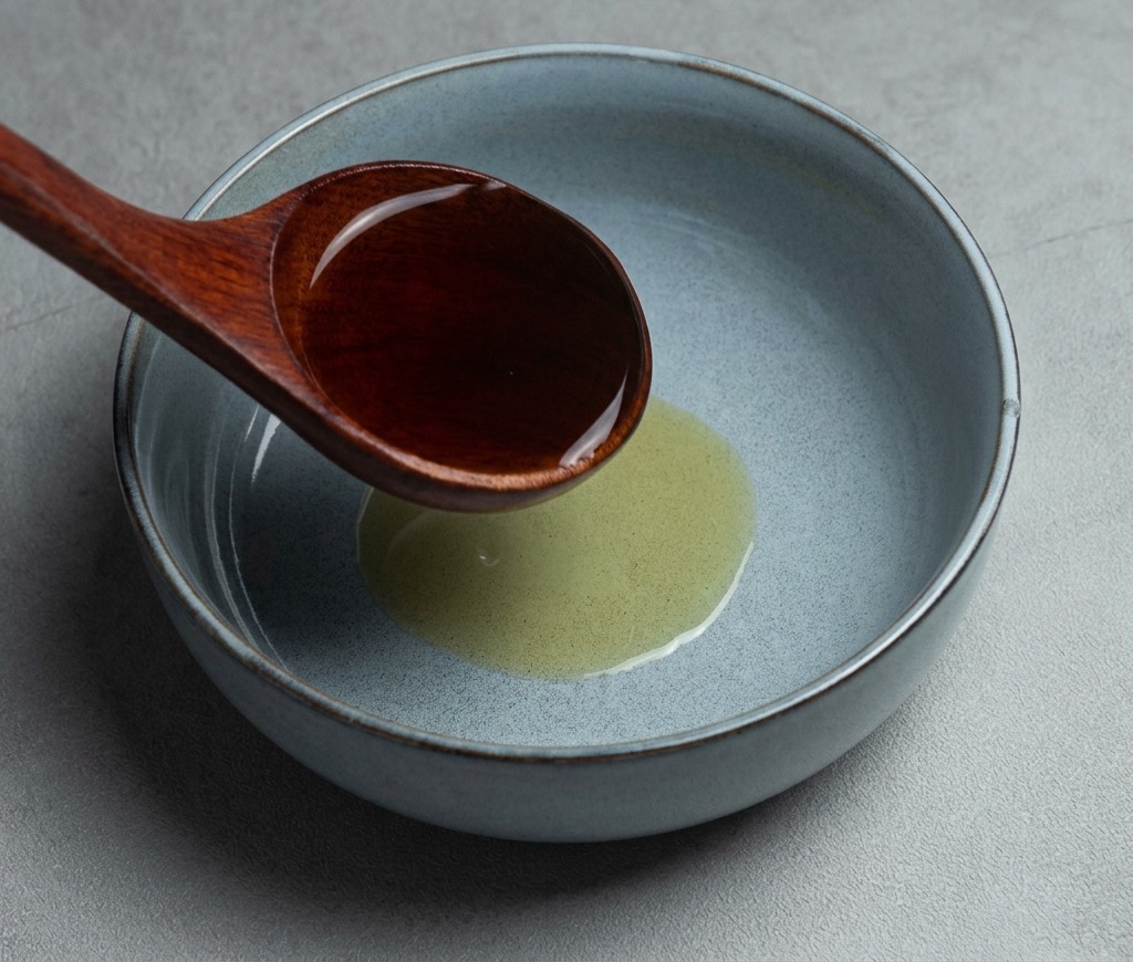 A dark wooden ladle pouring hot shoyu ramen broth into a gray ceramic bowl containing sesame oil.