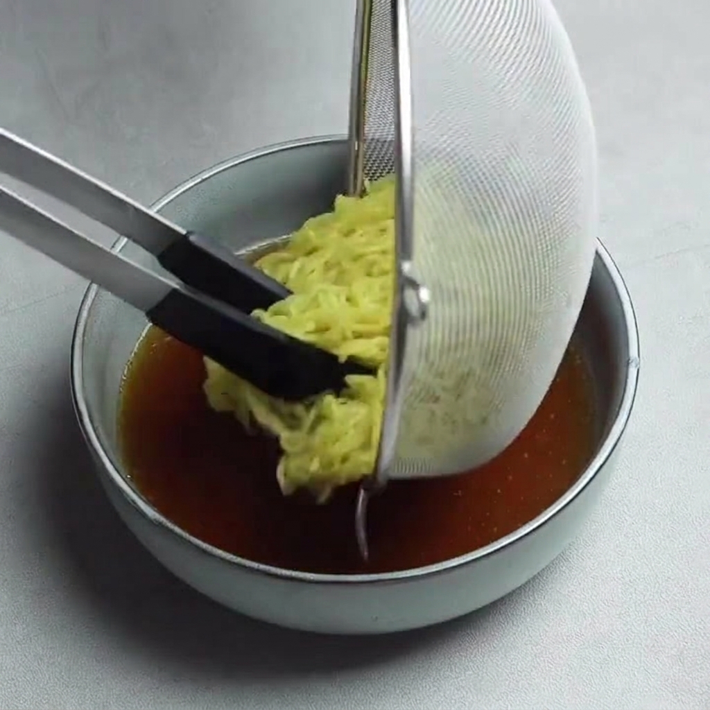 Yellow ramen noodles being lowered from a mesh strainer into a bowl of dark savory broth using black-handled tongs.