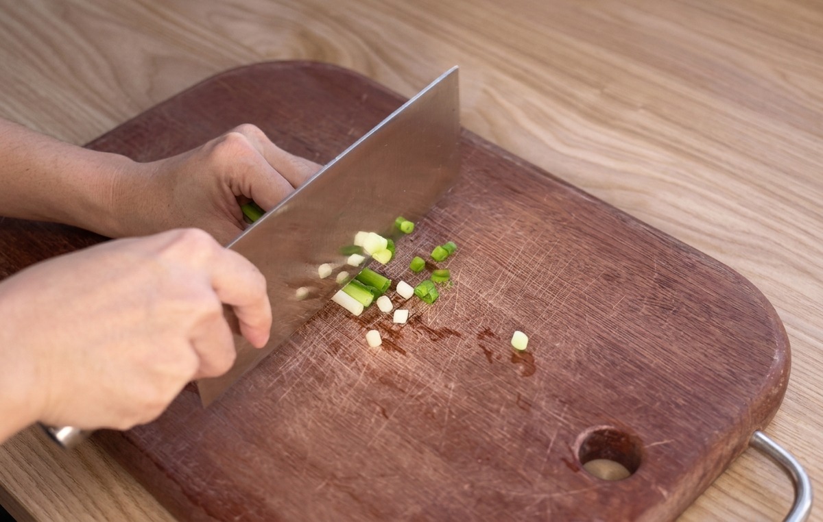 A persons hands using a large stainless steel cleaver to finely slice green onions on a worn wooden cutting board.