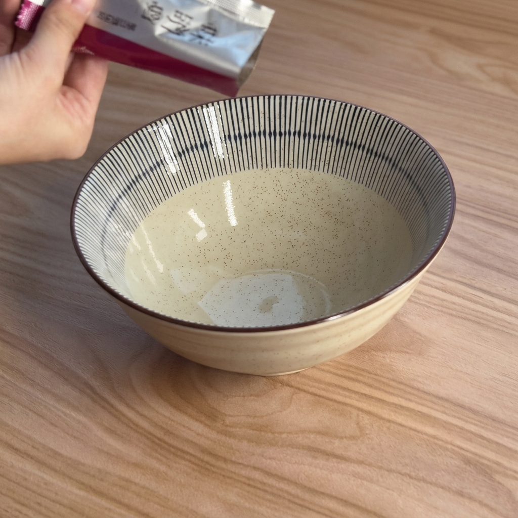 A hand squeezing a thick, brown miso paste from a silver foil packet into a cream-colored ceramic bowl with blue stripes.