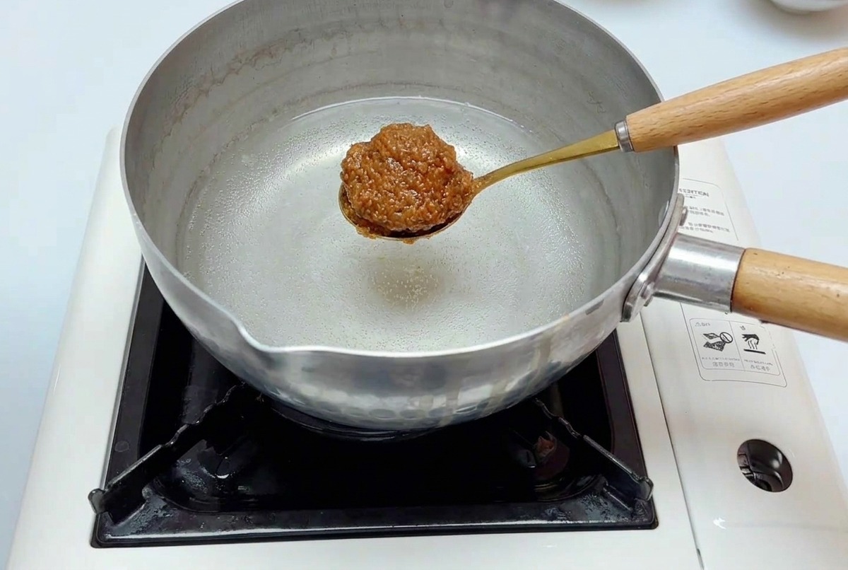 A golden spoon holding a portion of the miso mixture being lowered into a stainless steel pot of water on a stove.