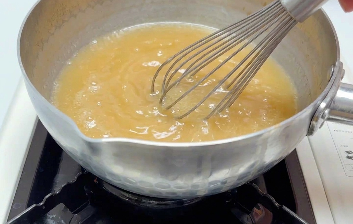 A hand whisking a cloudy, light-brown miso broth in a small saucepan over a lit burner.