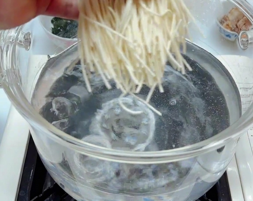 Fresh white ramen noodles being dropped into a clear glass pot of bubbling, boiling water.