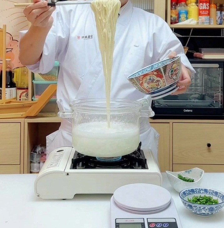 A portion of long, yellow ramen noodles being lifted high with chopsticks out of a steaming pot.