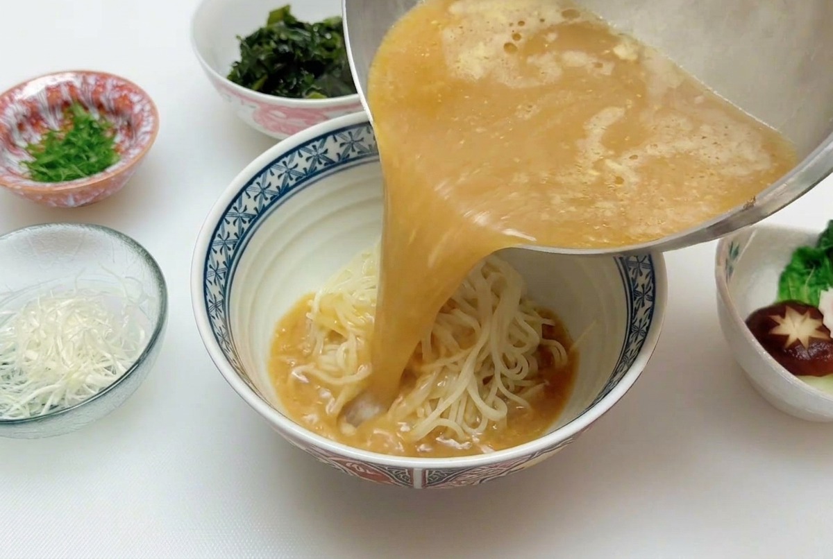 A steady stream of hot, golden-brown miso broth being poured from a saucepan onto a mound of ramen noodles in a bowl.