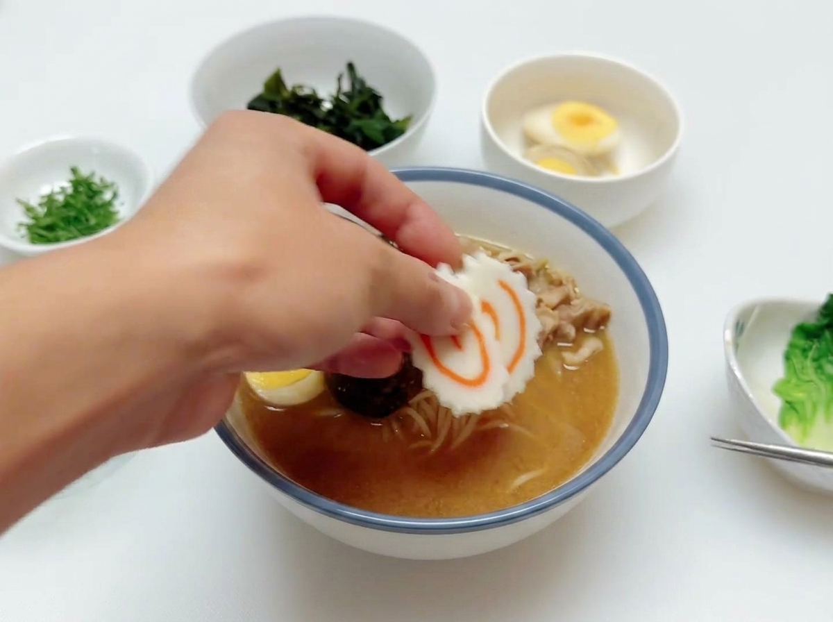 A hand placing a slice of pink and white swirled narutomaki fish cake onto a bowl of ramen already containing beef and egg.