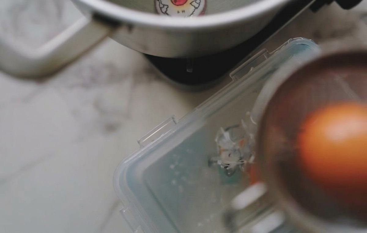 A brown egg being lowered into a clear container of ice and water using a metal slotted spoon.