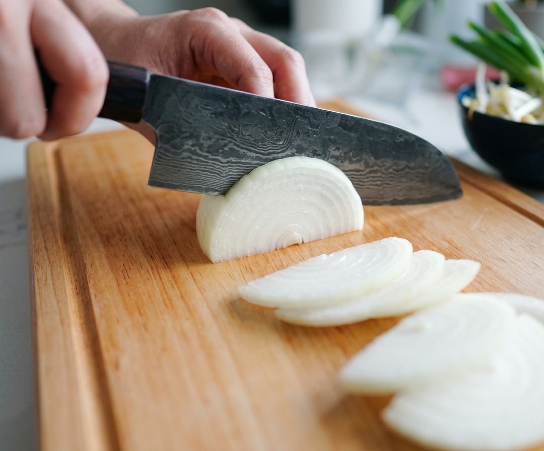 A person using a Damascus steel chefs knife to slice a white onion into thin half-moons on a wooden board.