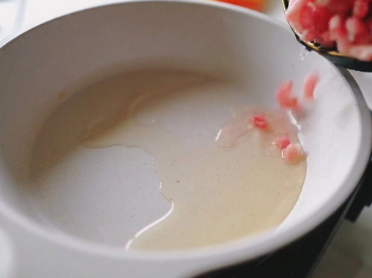 Fresh pink ground pork being added to a hot white frying pan with shimmering oil.