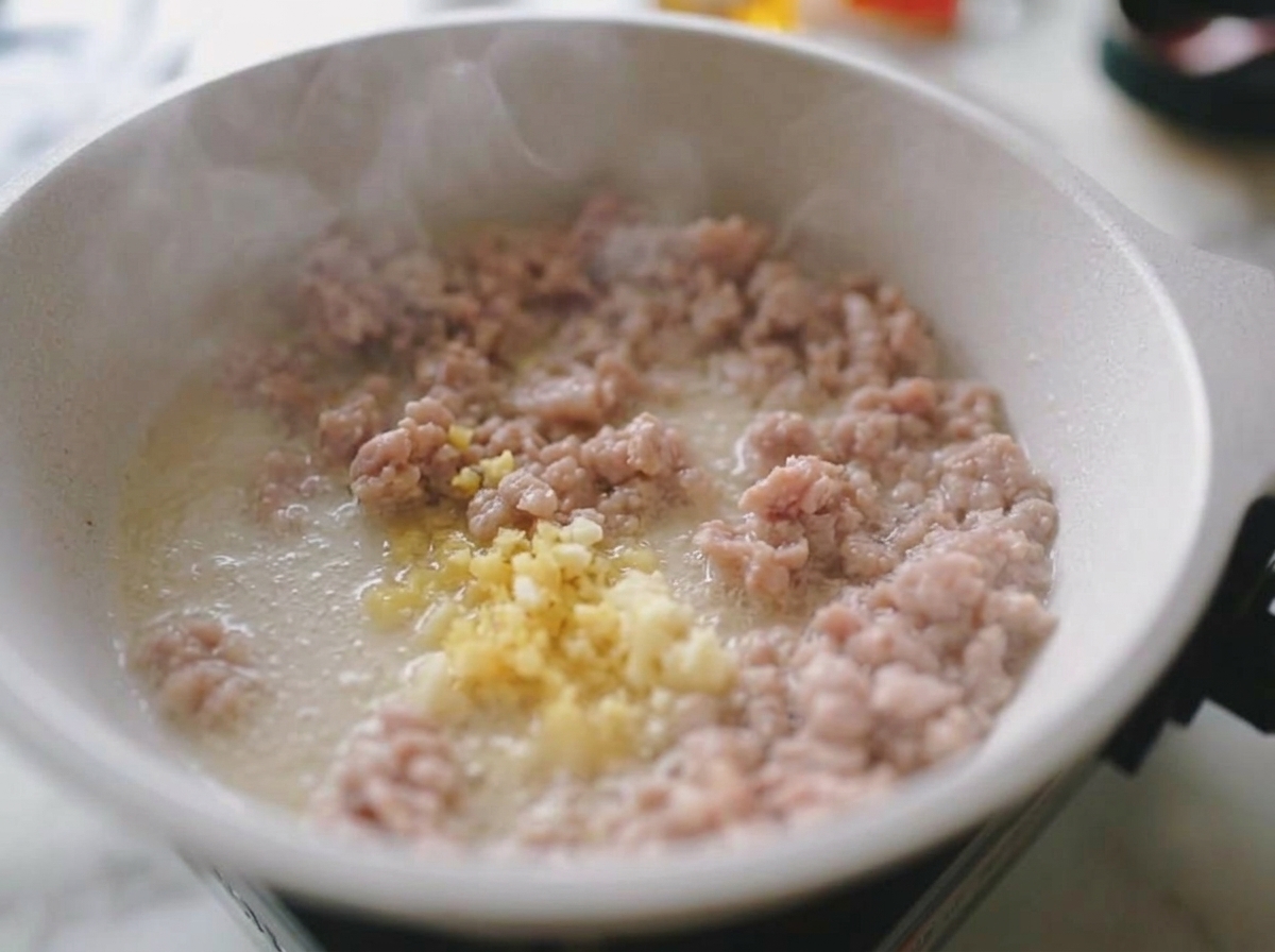 Minced garlic and ginger being stirred into browned ground pork in a white frying pan, with steam rising.