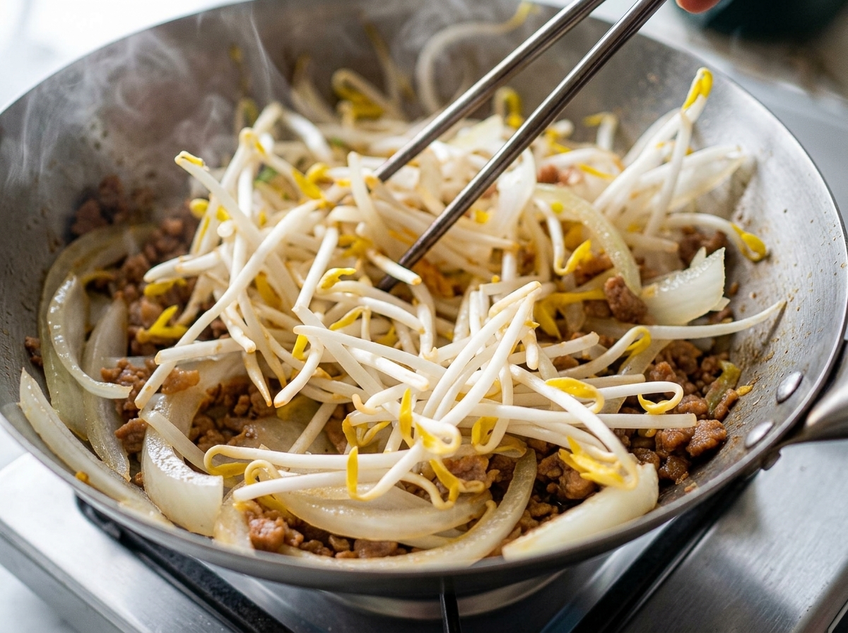 A large pile of white bean sprouts being tossed into a wok with browned ground meat and sliced onions.