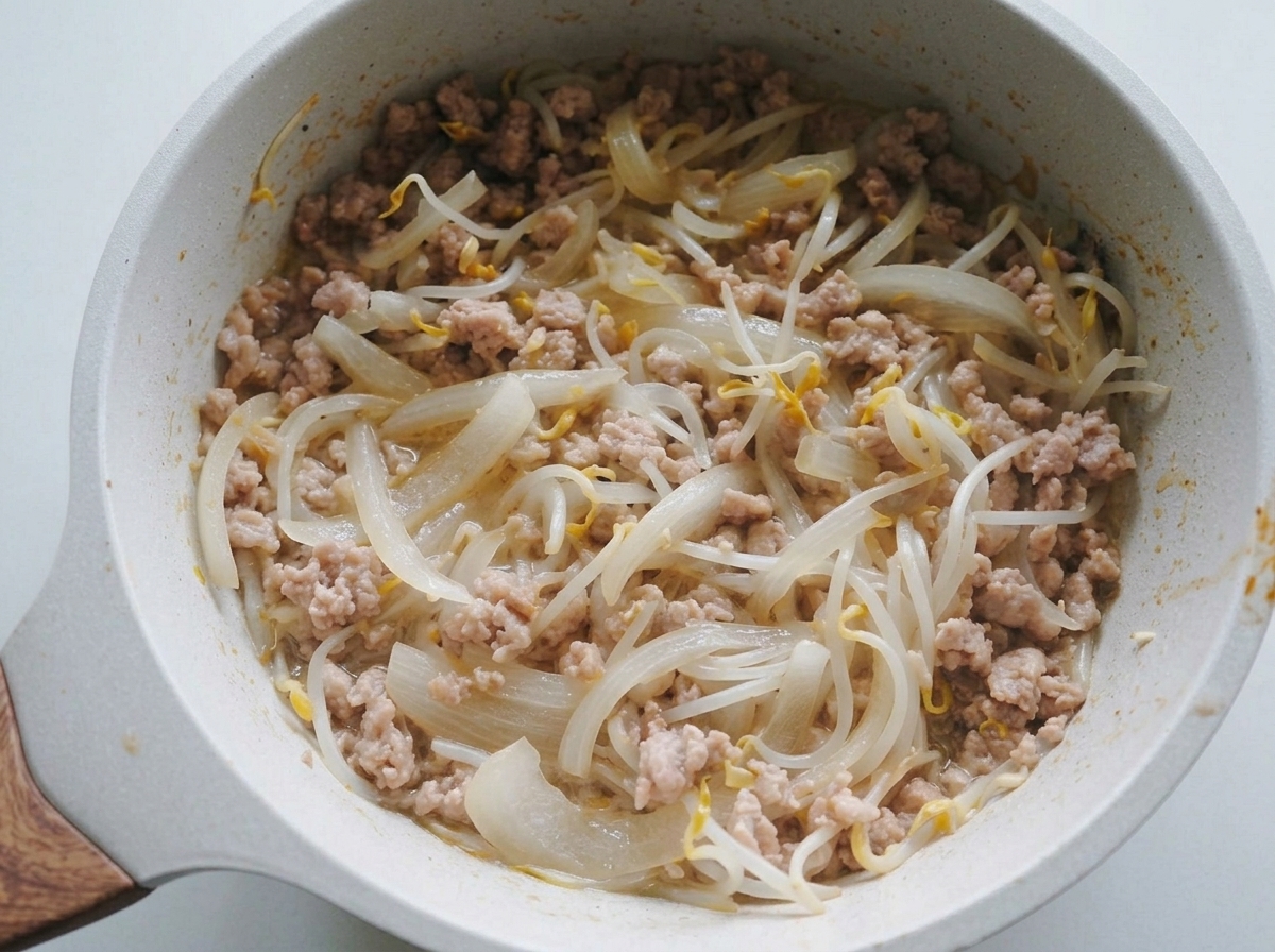 A mixture of sautéed onions, bean sprouts, and minced pork being stirred in a light-colored pan.