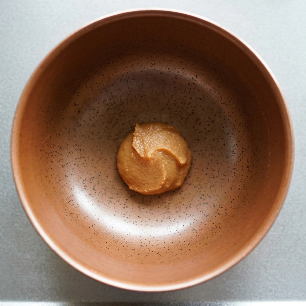 A dollop of thick brown miso paste sitting at the bottom of a speckled ceramic bowl.