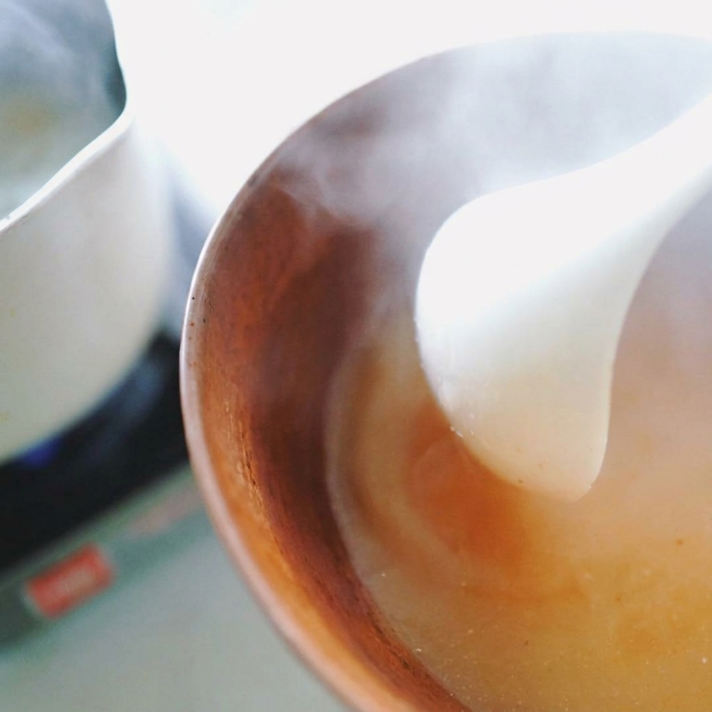 A white ladle stirring hot liquid into a brown bowl to dissolve miso paste into a steaming broth.