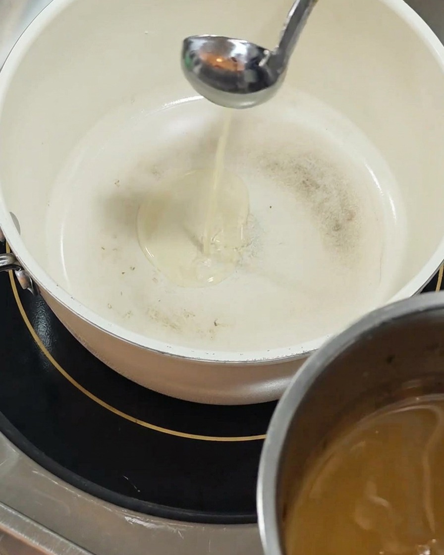 A silver ladle pours clear yellow oil into a white ceramic-coated pot sitting on a black induction burner.
