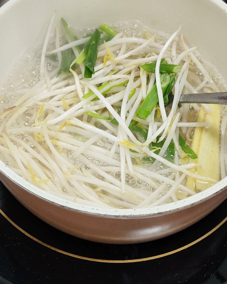 A pot of simmering water containing bean sprouts, chopped green garlic leaves, and pieces of baby corn being stirred with a metal utensil.