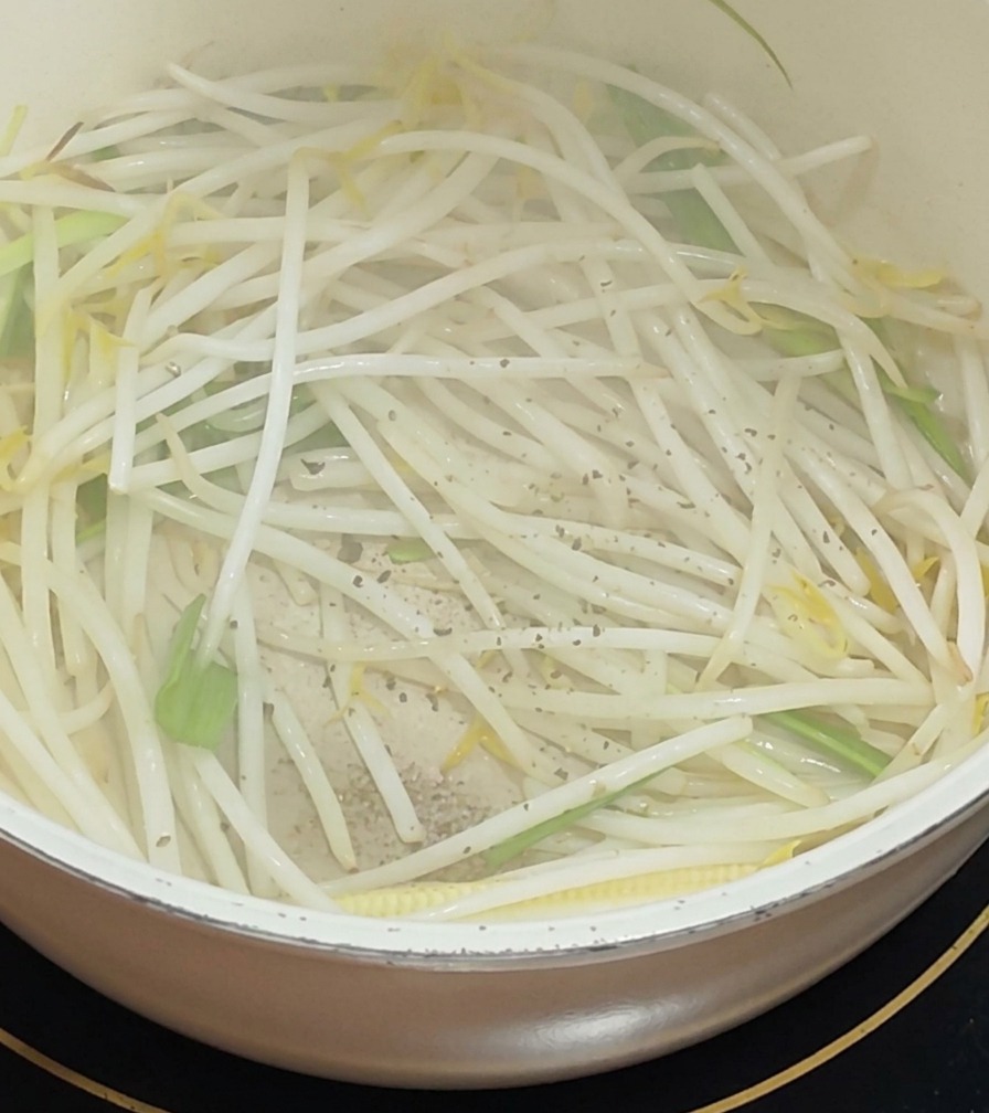 Black pepper being ground over a white pot filled with sautéed bean sprouts and green scallion pieces.