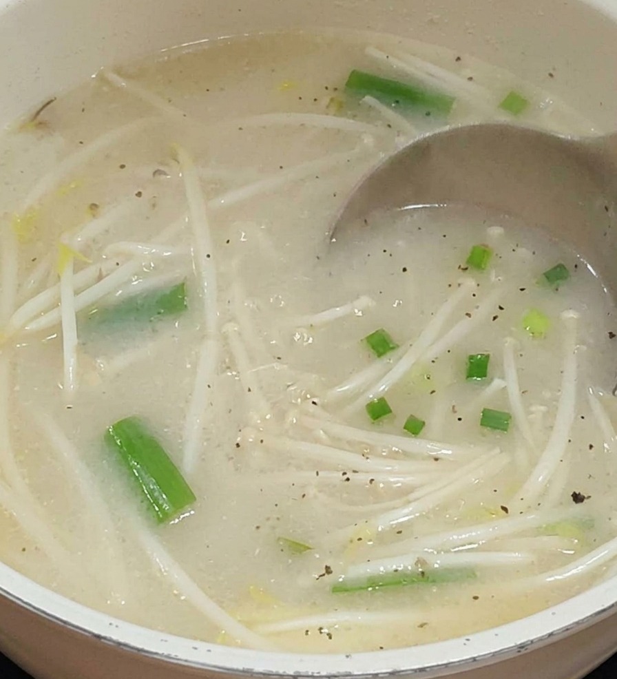 A metal ladle pouring a milky, light-colored broth into a pot containing bean sprouts and green vegetables.