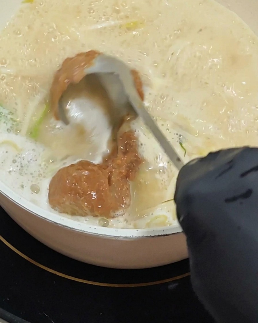 A gloved hand using a ladle to stir a thick scoop of reddish-brown miso paste into a bubbling, creamy soup base.