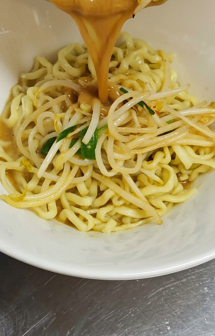 A stream of thick, brown miso broth and sautéed bean sprouts being poured from a pan into a white bowl filled with wavy yellow ramen noodles.