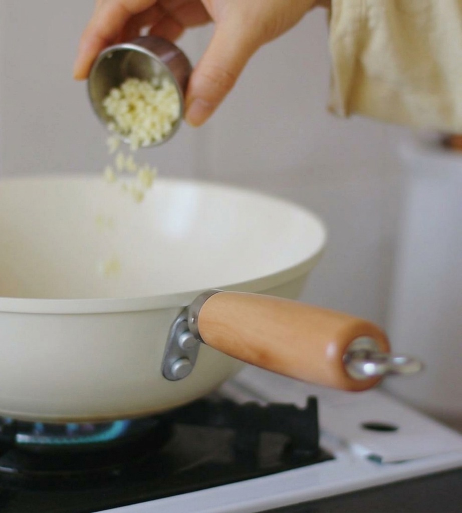A hand pouring finely minced garlic from a small stainless steel bowl into a cream-colored pan resting on a gas stove with a blue flame.