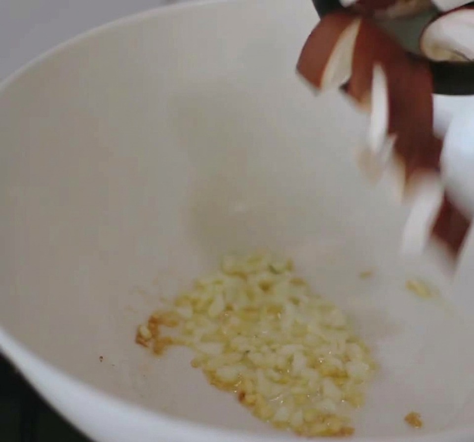 A white pan on a stove containing sautéed minced garlic with sliced shiitake mushrooms being added from above.