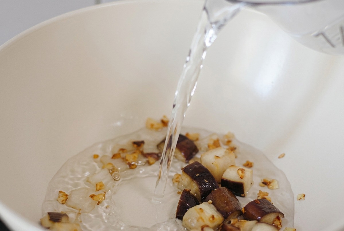 Water being poured from a pitcher into a white pan containing pieces of sautéed mushroom and garlic.
