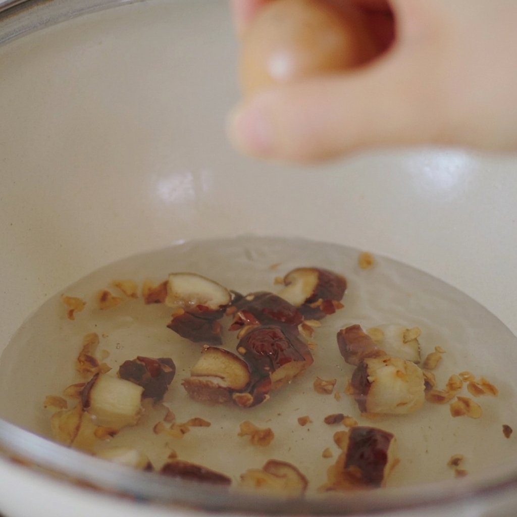 A hand lowering a brown chicken egg into a white pot filled with water and sliced shiitake mushrooms.