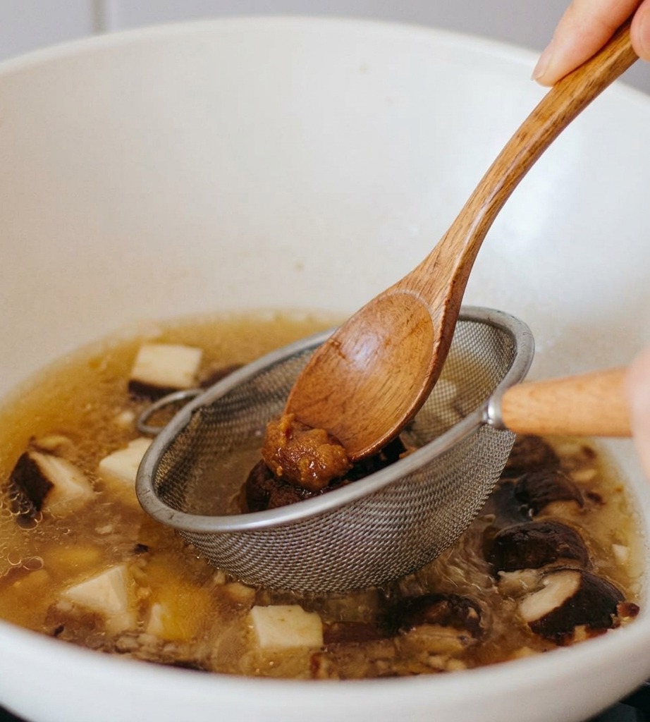 A wooden spoon pressing dark miso paste through a small metal strainer into a simmering broth with mushroom slices.