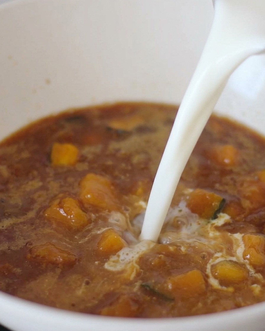 A stream of white milk being poured into a dark brown miso broth containing pieces of vegetables.