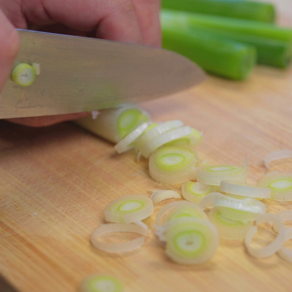A close-up shot of a chefs knife slicing through a bunch of fresh green onions on a light wooden cutting board.