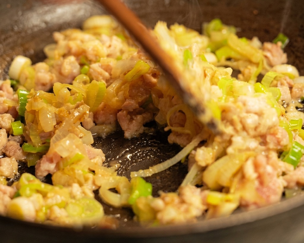 A wooden spatula stir-frying browned ground pork and green onions in a hot pan with steam rising.