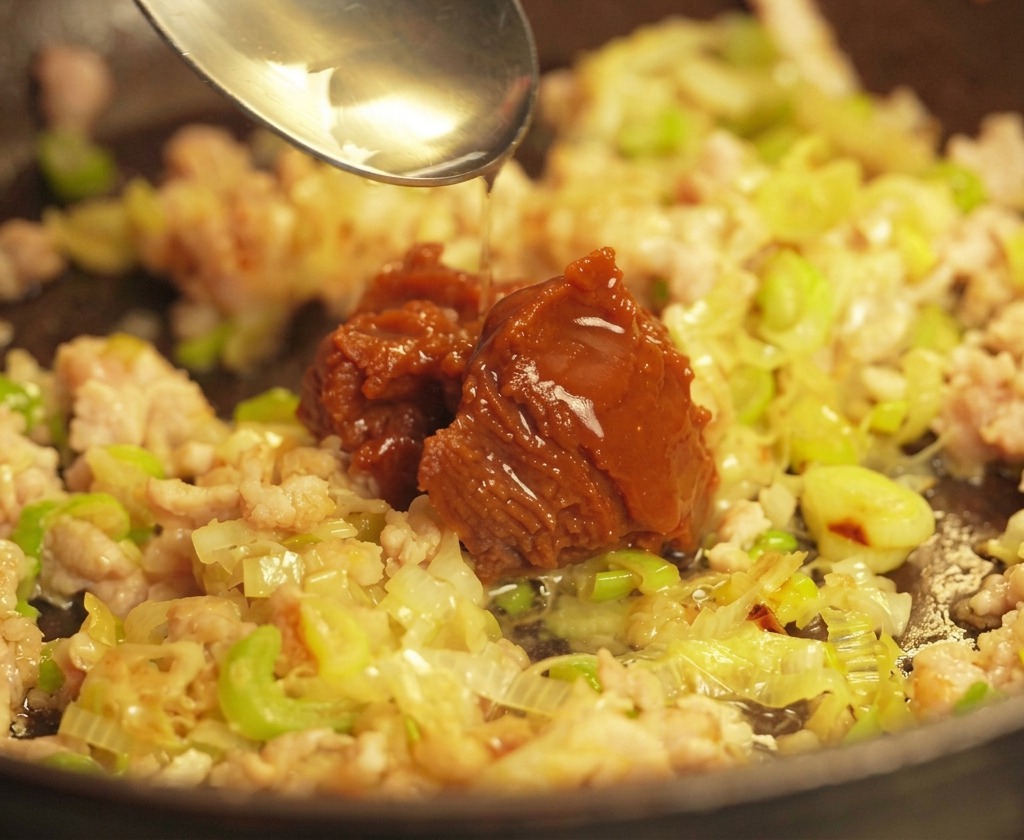A silver spoon dropping a large dollop of dark brown miso paste onto a bed of sautéed ground pork and onions in a pan.