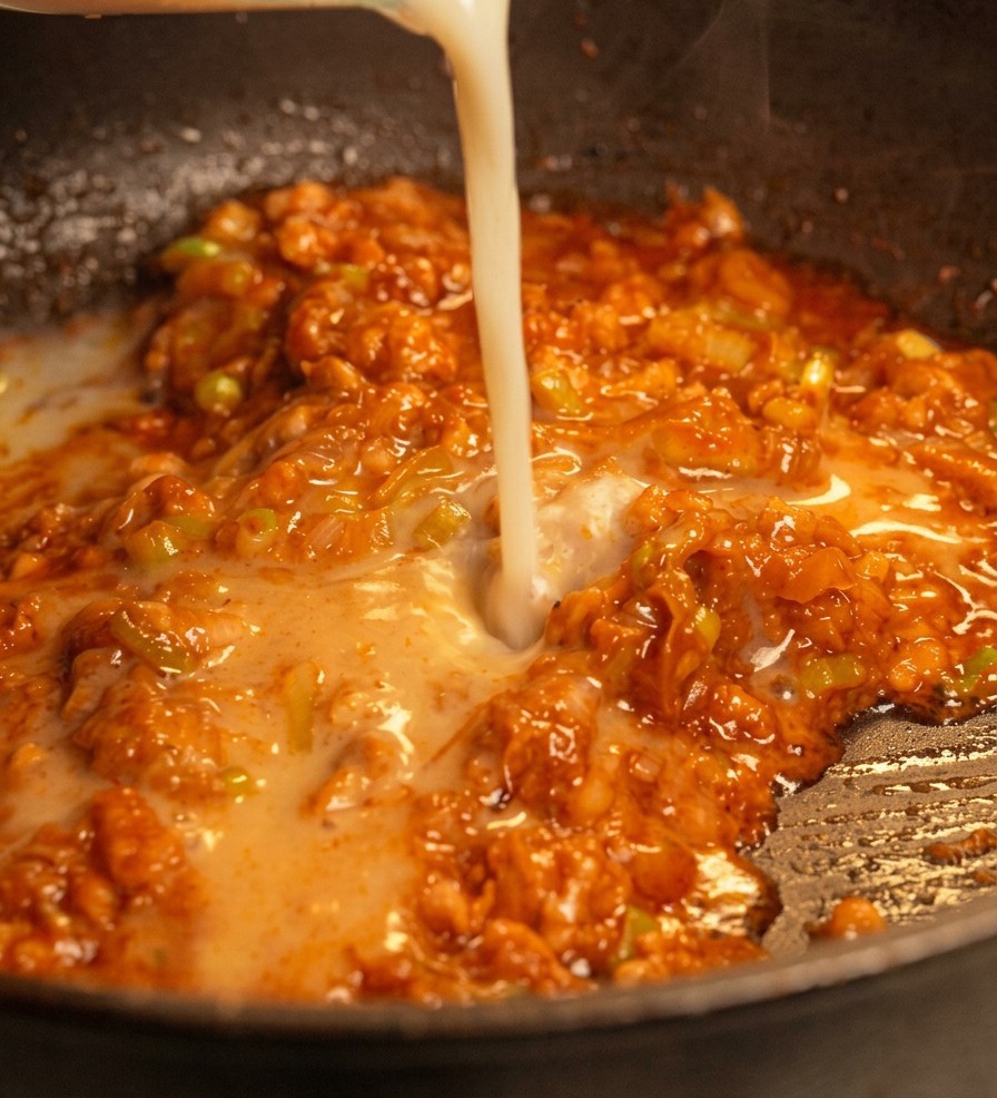 A stream of creamy white soy milk being poured into a pan containing a red-tinted, savory pork and miso mixture.