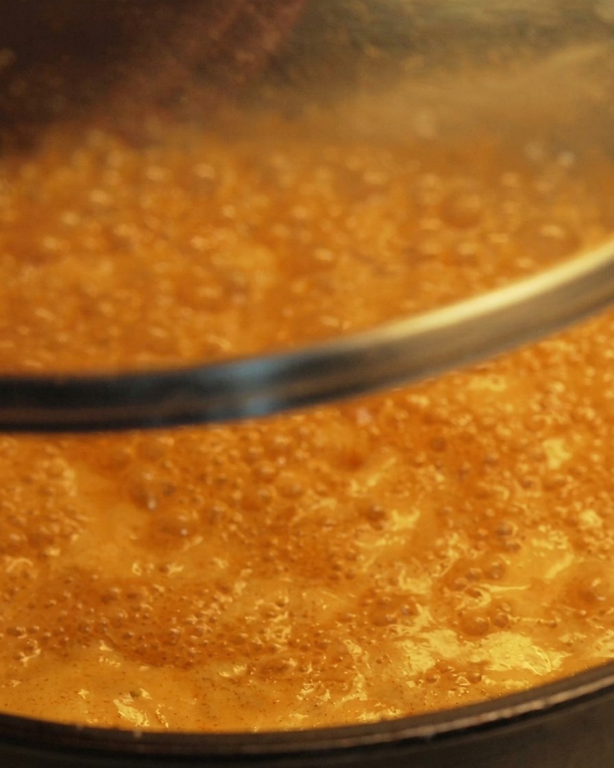 A clear glass lid being lowered onto a pan containing a bubbling, simmering orange-colored sauce.