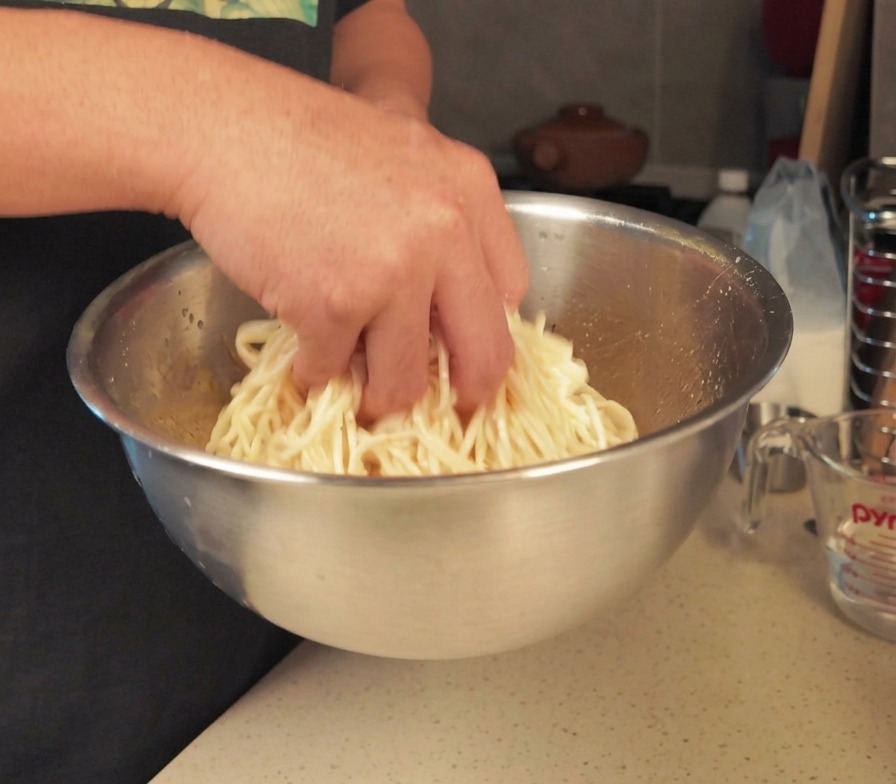A person mixing a batch of cooked white noodles with their hand in a large stainless steel bowl to coat them with oil.