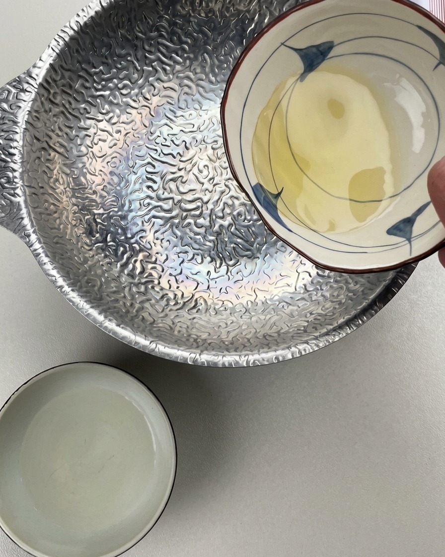 A person pouring mirin from a small ceramic bowl into a silver textured pot, with another bowl of sake waiting on the counter.