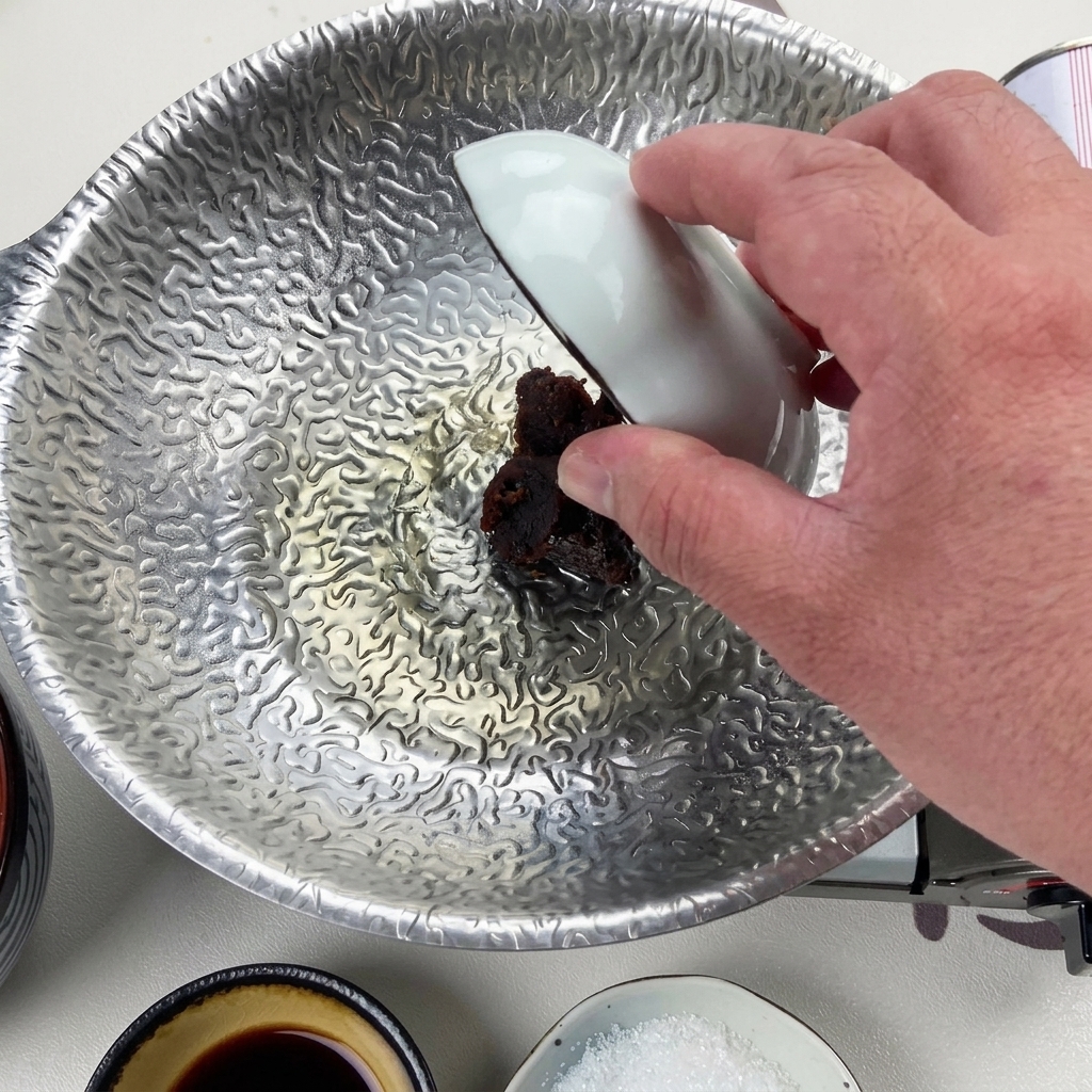 A hand using a small white bowl to drop a thick scoop of dark red miso paste into the reduced liquid at the bottom of the silver pot.